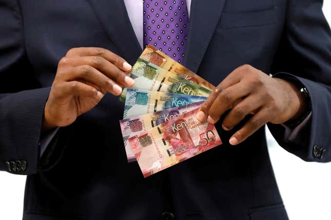 A person in a dark suit and purple tie holds colorful Kenyan bank notes. 