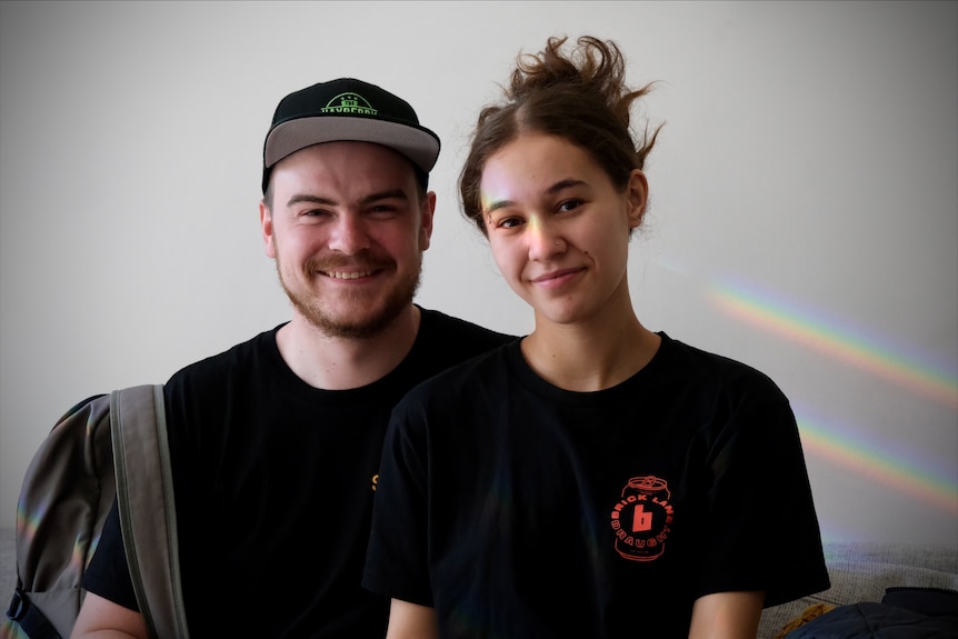 A man and woman in their mid-20s. The man is wearing a cap and has a beard, and the woman has her long brown hair tied up.