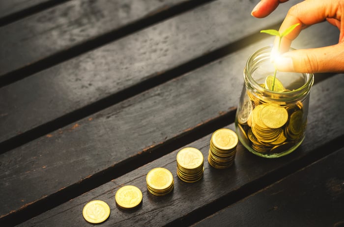 Hand touching sapling in jar of gold coins next to increasingly taller stacks of coins.