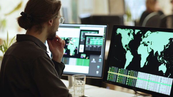 Person wearing glasses is surrounded by computer monitors in an office. In front of him there are screens showing maps of the world with associated data.