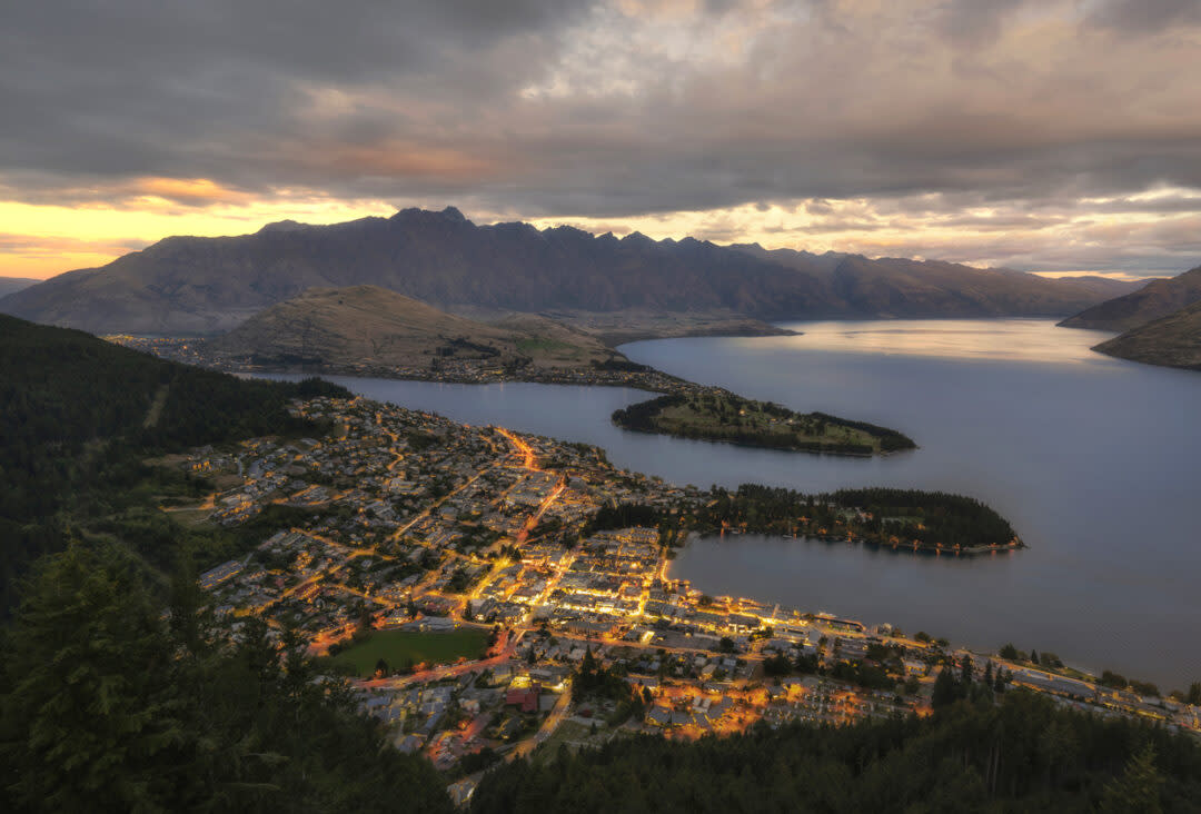 Aerial view of a town beside a lake under a cloudy, dramatic sunset sky. The town is illuminated with lights, nestled between lush greenery and mountains. A large body of water reflects the evening light.