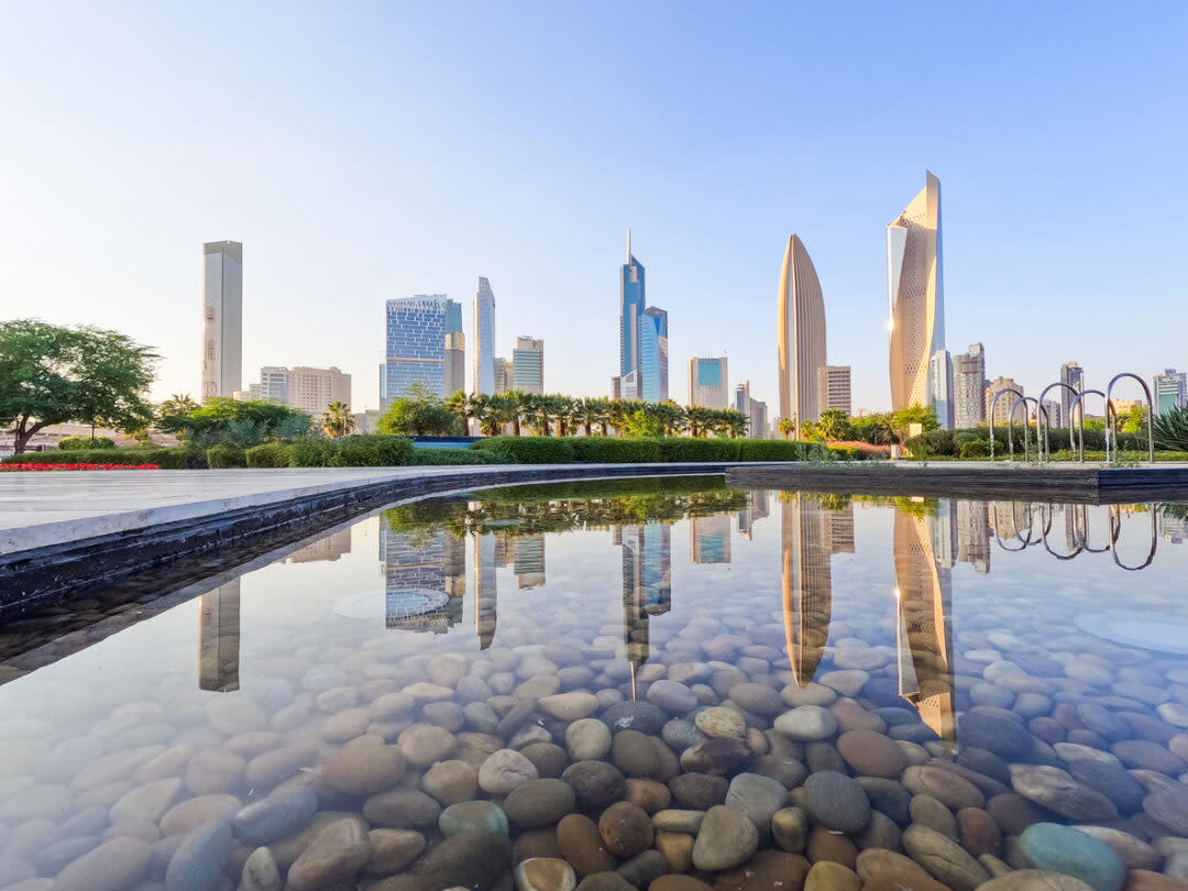 Skyline of modern skyscrapers in a city reflected in a clear water pool with smooth stones at the bottom. The scene is set under a clear blue sky, with trees and greenery visible in the background.