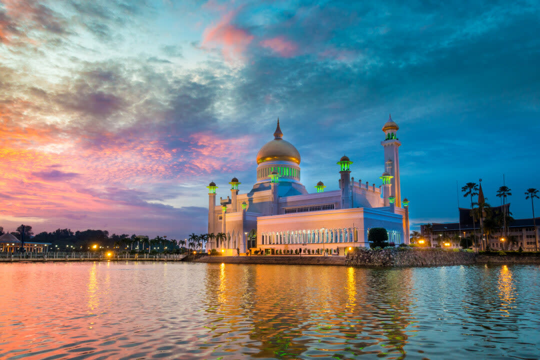 Golden-domed mosque with minarets by a waterfront at sunset. The sky displays hues of orange, pink, and blue, reflecting on the water. Palm trees line the shore, and the mosque is illuminated, enhancing its majestic appearance.