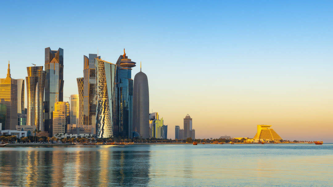 Skyline of Doha, Qatar, showcasing modern skyscrapers and unique architecture along the waterfront. The sun sets, casting a warm glow over the buildings and calm waters, with a clear blue sky above.