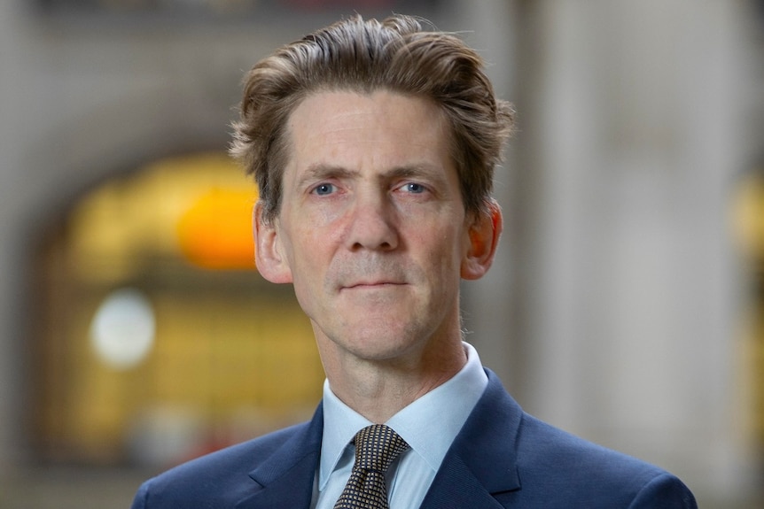 An older man wearing a blue suit, light blue collared shirt and patterned tie in front of a building with a steely expression.