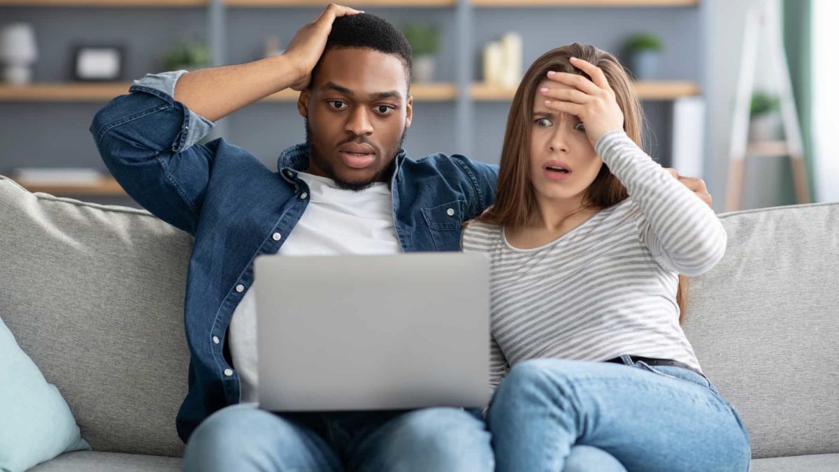 A couple sits on a sofa, each clutching their heads in horror and disbelief, while looking at a laptop screen.