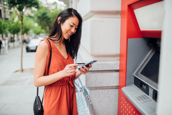 Person holding a banking card and smartphone in their hands while standing at an ATM.