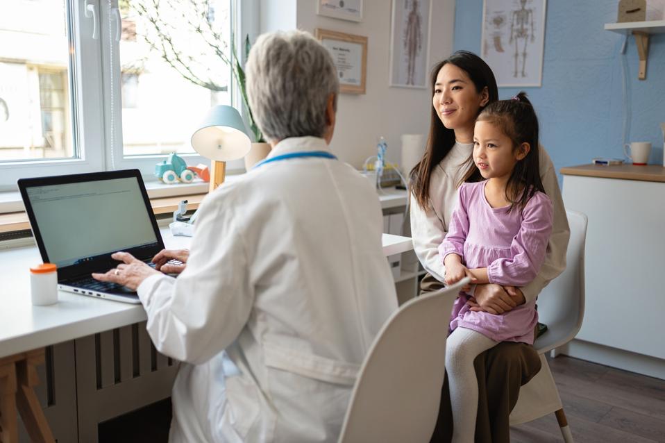 Senior Caucasian female pediatrician enters Japanese girl's information into laptop, during annual medical check-up