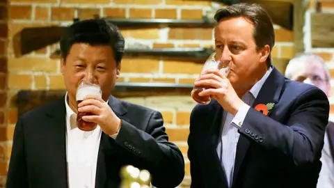 Getty Images Xi Jinping and David Cameron wearing suits and open-necked shirts, holding and drinking pints of beer in a rustic setting with exposed brick walls and mounted antique rifles in the background.
