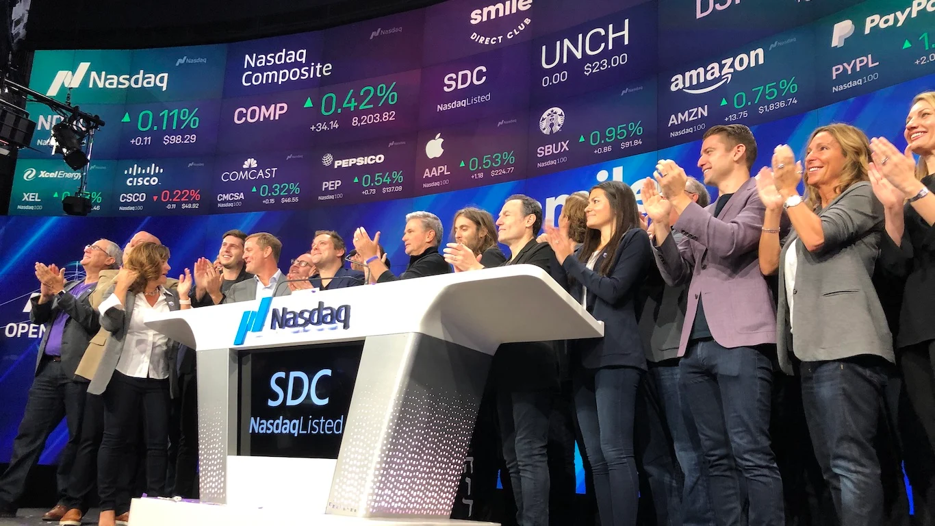 A group of diverse individuals, including men and women in business attire, are clapping enthusiastically behind a Nasdaq branded podium. Behind them, a large digital display shows various stock market information, including Nasdaq Composite, Amazon, Apple, Cisco, PepsiCo, Starbucks, and others, with their respective stock symbols, percentages, and prices. The screen also features Nasdaq logos prominently.