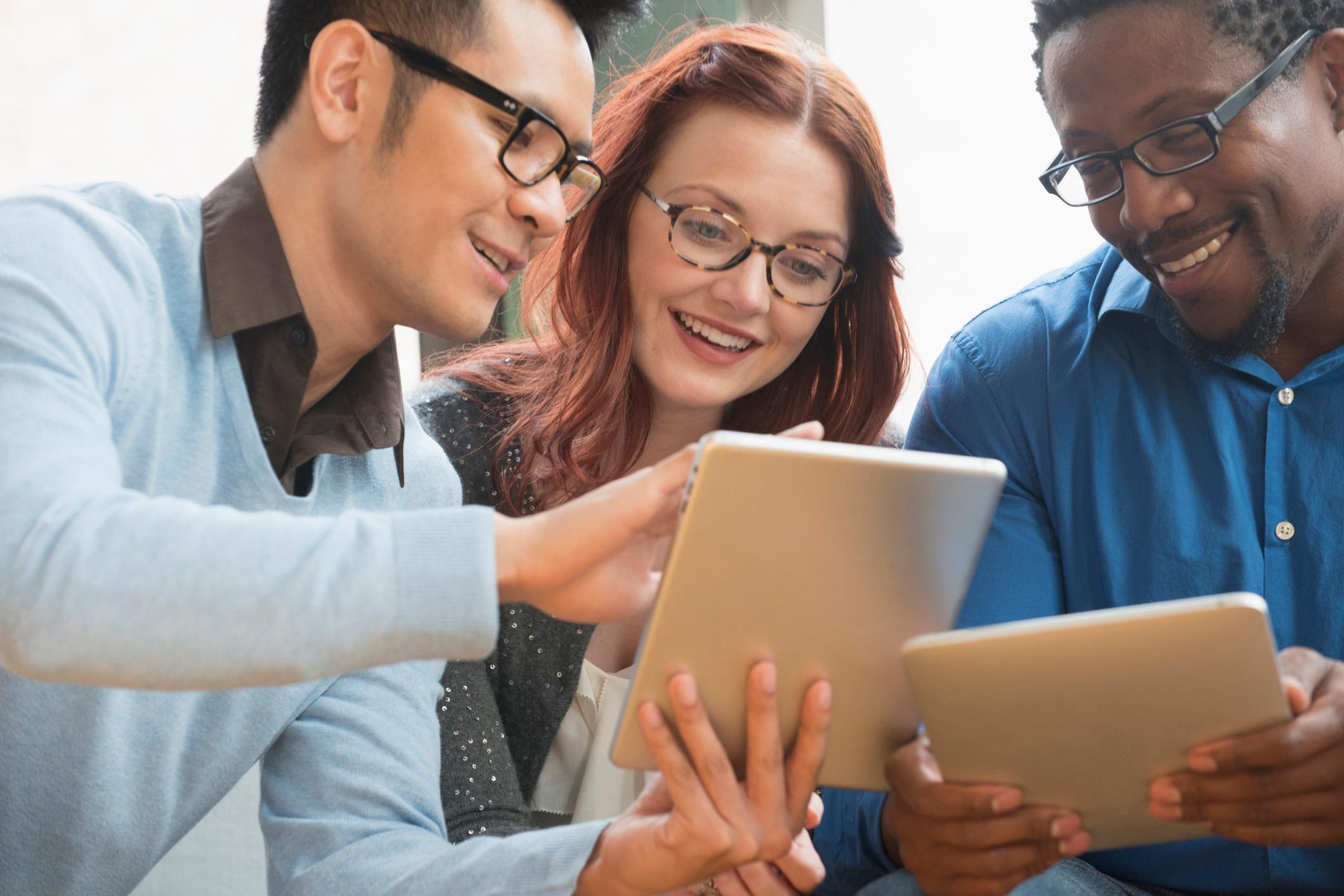 Three smiling colleagues collaborate while viewing tablets in a bright office.