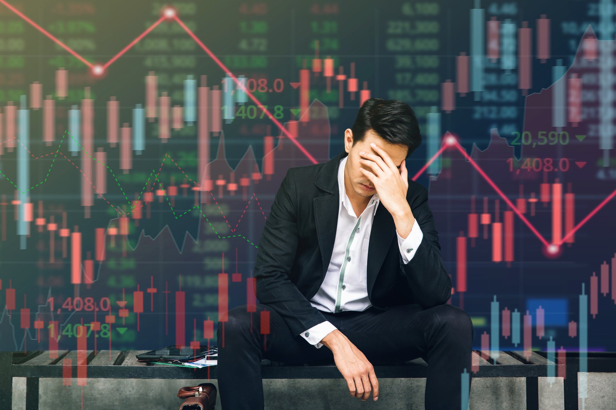 Person sitting with hand over face in front of large stock chart showing red data points.