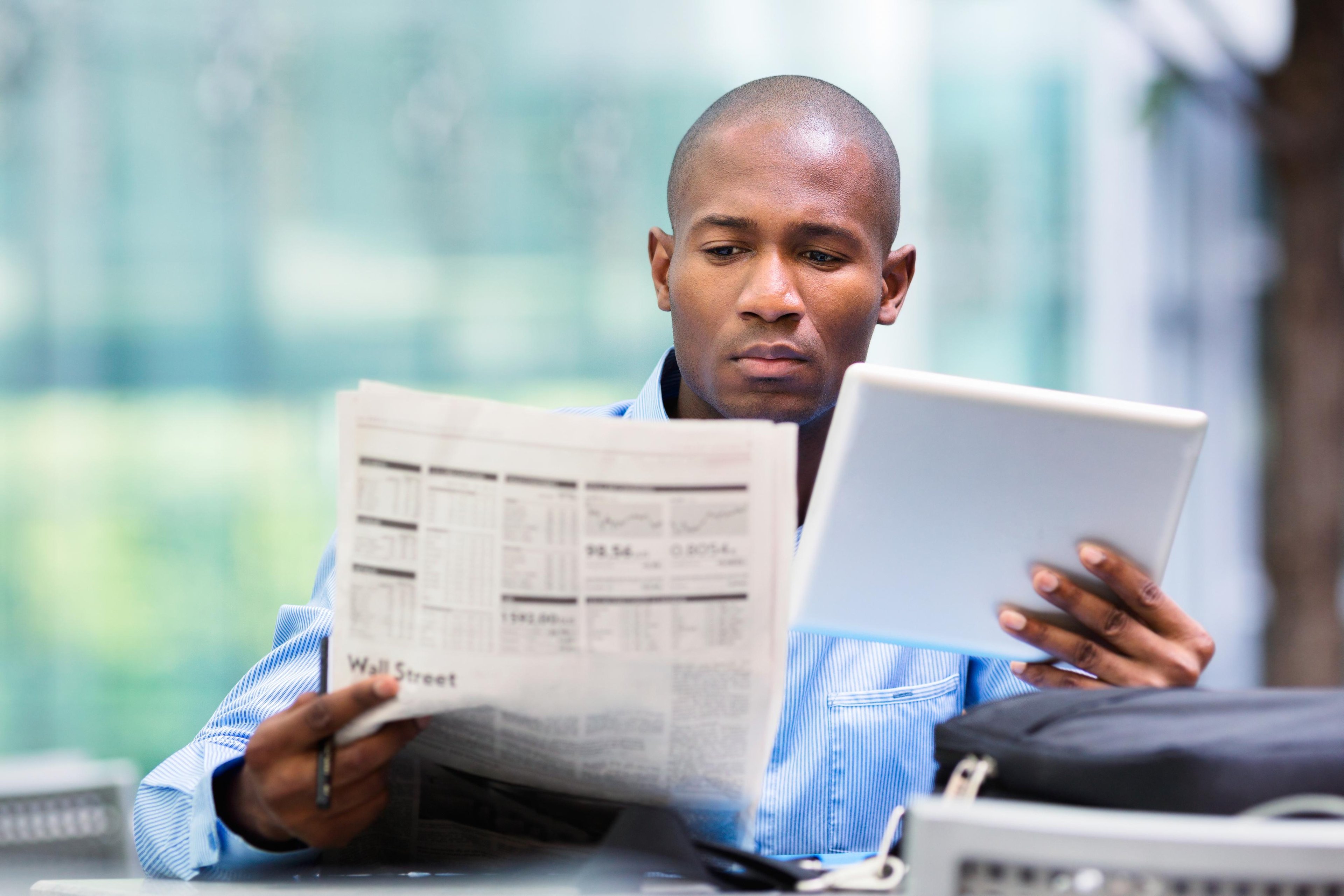 A seated businessperson is reading a financial newspaper while holding a tablet in their left hand.