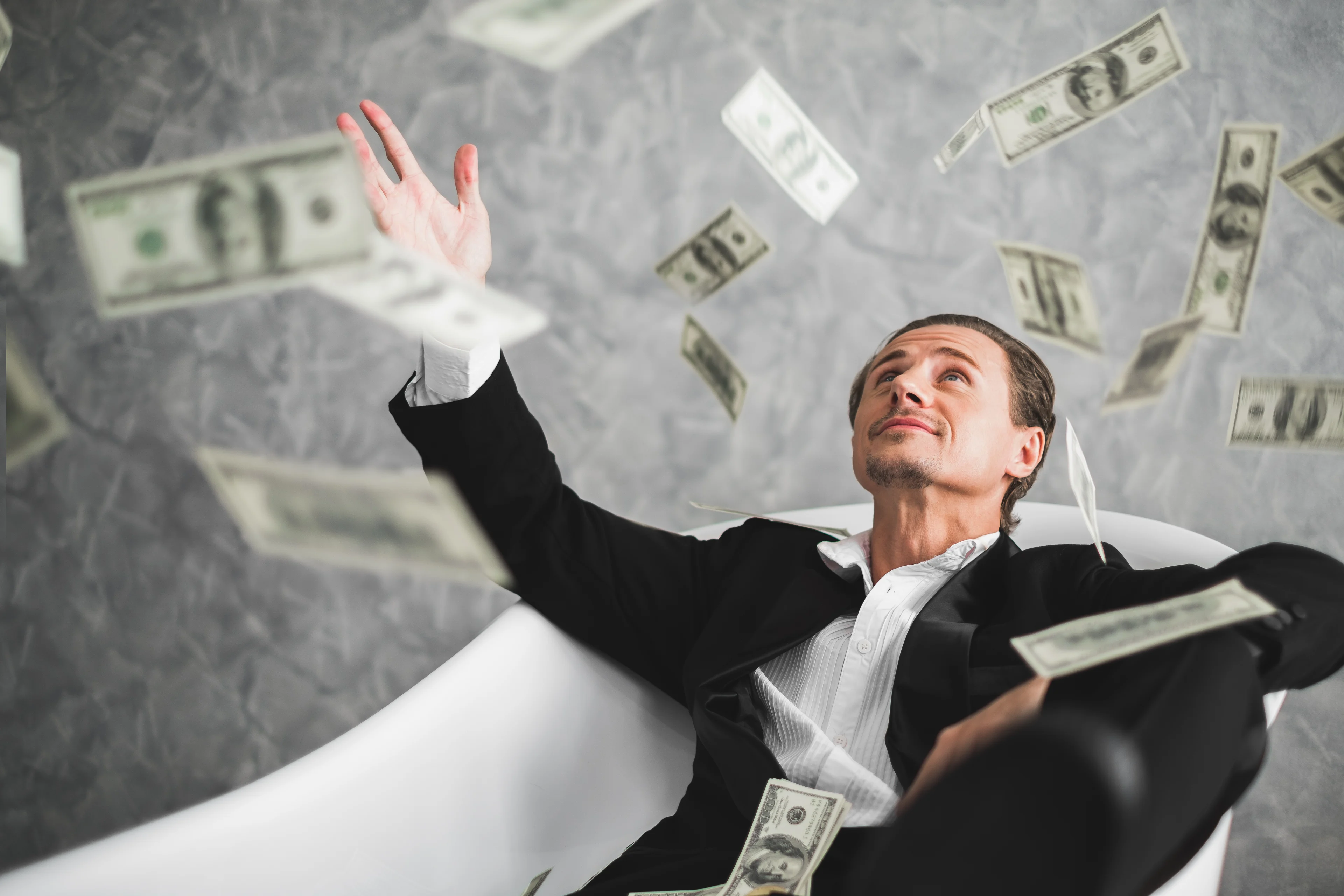Man in a suit sitting in a bathtub amid flying currency notes.